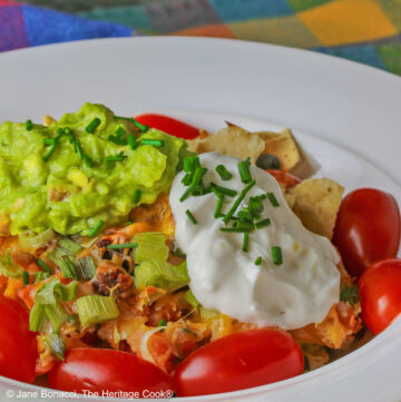 A serving of Mexican Chilaquiles Casserole, made of layers of tortilla chips, salsa, beans, green onions, cheese, and chorizo in a bowl with a wide white brim with dollops of guacamole and sour cream on top, cherry tomatoes around the edges, and chives sprinkled over the top; bowl is sitting on brightly colored cloths © 2025 Jane Bonacci, The Heritage Cook.