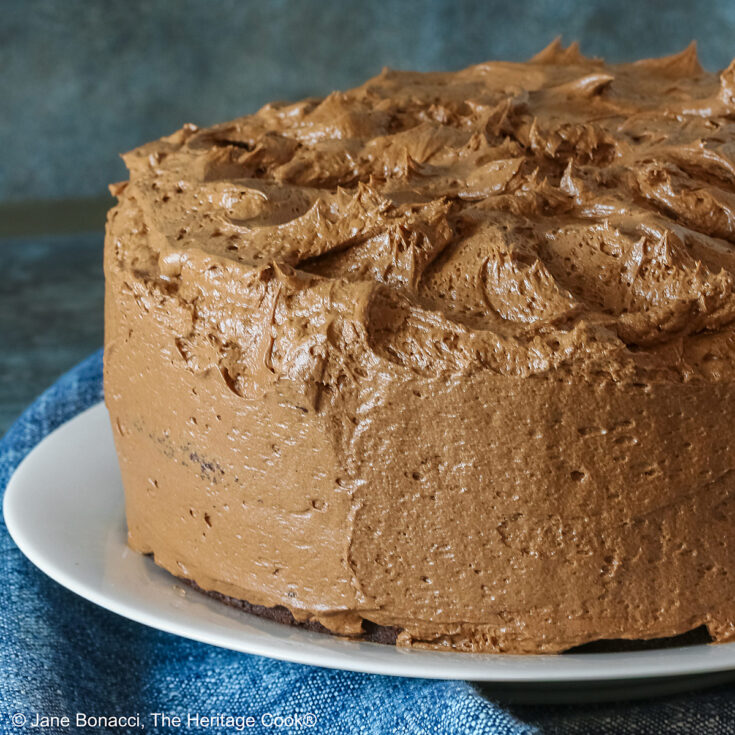 Large whole chocolate mocha layer cake with chocolate buttercream frosting, on a white plate sitting on a blue cloth, some with a cut slice on a white plate with a fork in front of the whole cake © 2025 Jane Bonacci, The Heritage Cook.
