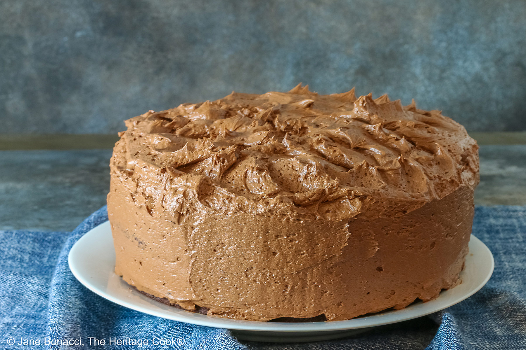 Large whole chocolate mocha layer cake with chocolate buttercream frosting, on a white plate sitting on a blue cloth, some with a cut slice on a white plate with a fork in front of the whole cake © 2025 Jane Bonacci, The Heritage Cook. 