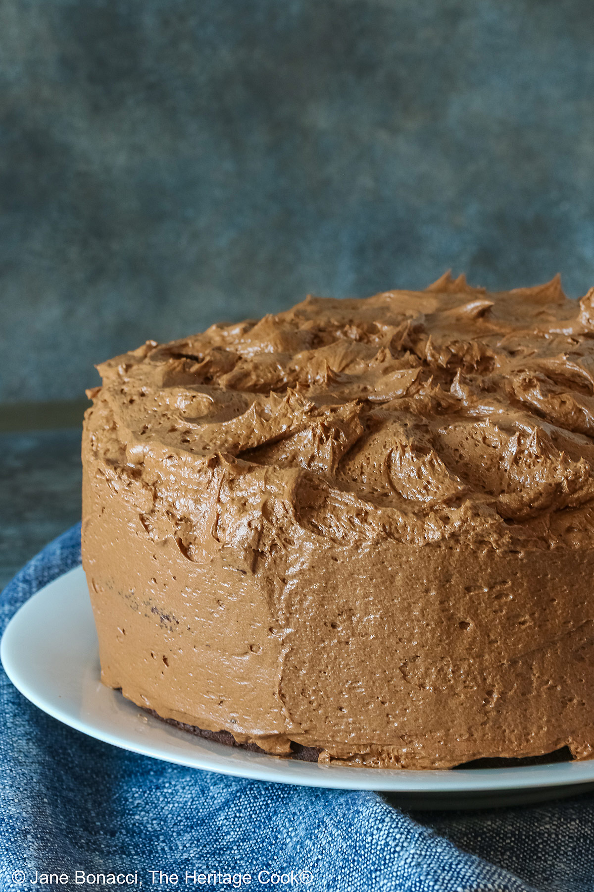 Large whole chocolate mocha layer cake with chocolate buttercream frosting, on a white plate sitting on a blue cloth, some with a cut slice on a white plate with a fork in front of the whole cake © 2025 Jane Bonacci, The Heritage Cook. 