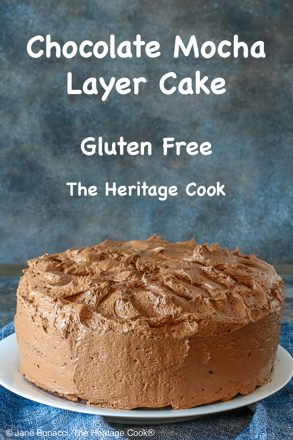 Large whole chocolate mocha layer cake with chocolate buttercream frosting, on a white plate sitting on a blue cloth, some with a cut slice on a white plate with a fork in front of the whole cake © 2025 Jane Bonacci, The Heritage Cook. 