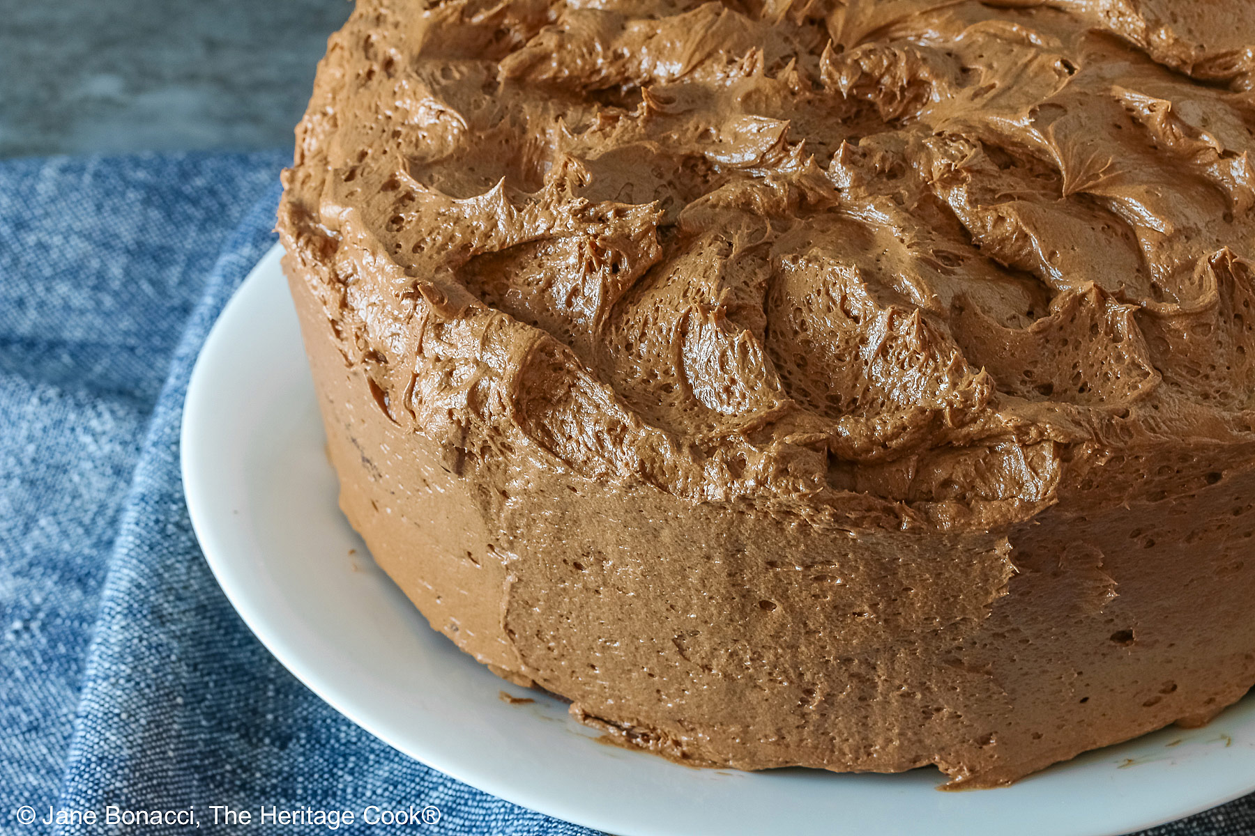 Large whole chocolate mocha layer cake with chocolate buttercream frosting, on a white plate sitting on a blue cloth, some with a cut slice on a white plate with a fork in front of the whole cake © 2025 Jane Bonacci, The Heritage Cook. 