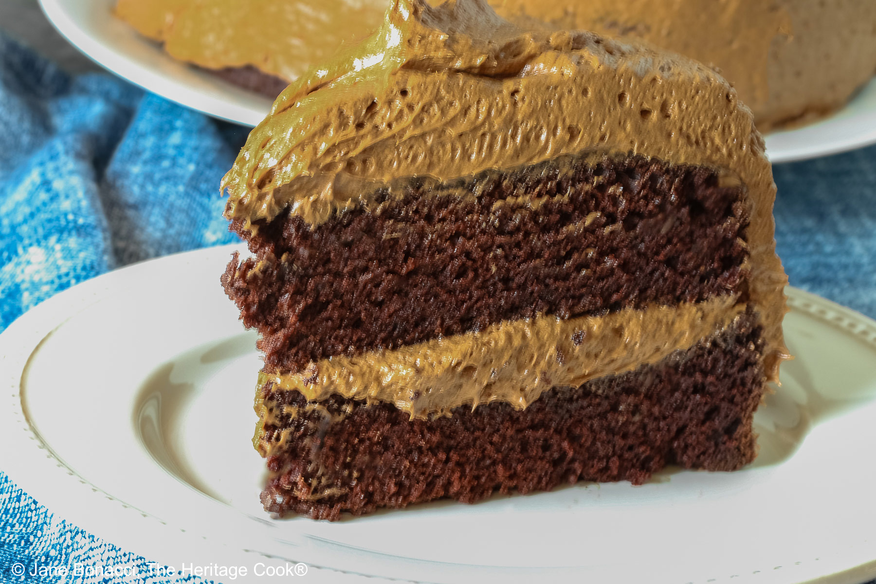 A slice of the chocolate mocha layer cake with chocolate frosting on a white plate sitting on a blue cloth. 