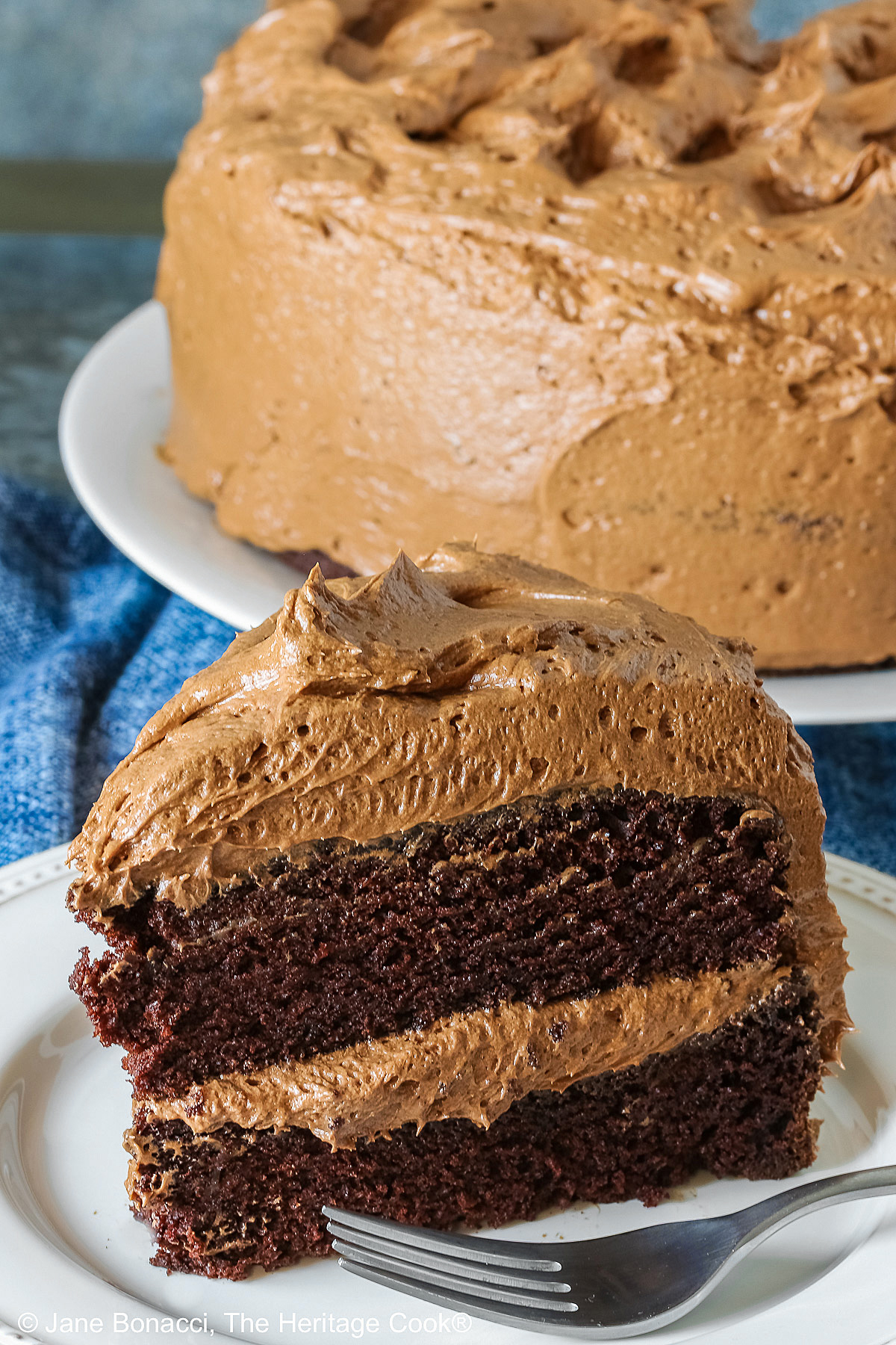 Large whole chocolate mocha layer cake with chocolate buttercream frosting, on a white plate sitting on a blue cloth, some with a cut slice on a white plate with a fork in front of the whole cake © 2025 Jane Bonacci, The Heritage Cook. 