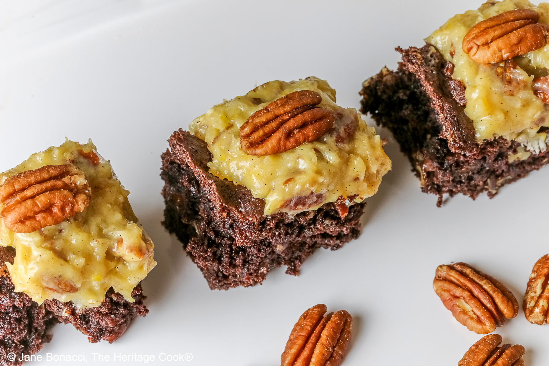 Dark brownies topped with golden pecan/coconut frosting, three squares in a row on a white rectangular tray, sitting on a blue cloth with pecan halves in front © 2025 Jane Bonacci, The Heritage Cook.