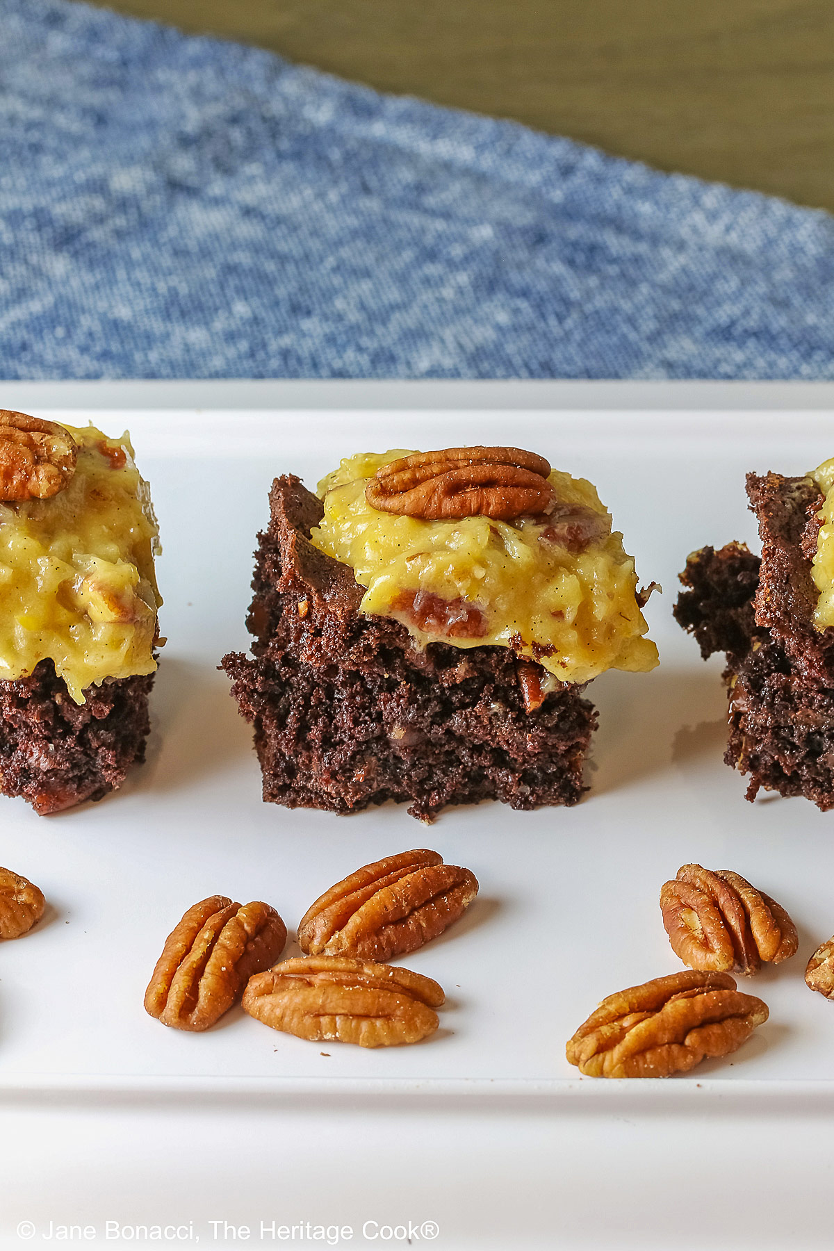 Dark brownies topped with golden pecan/coconut frosting, three squares in a row on a white rectangular tray, sitting on a blue cloth with pecan halves in front © 2025 Jane Bonacci, The Heritage Cook.