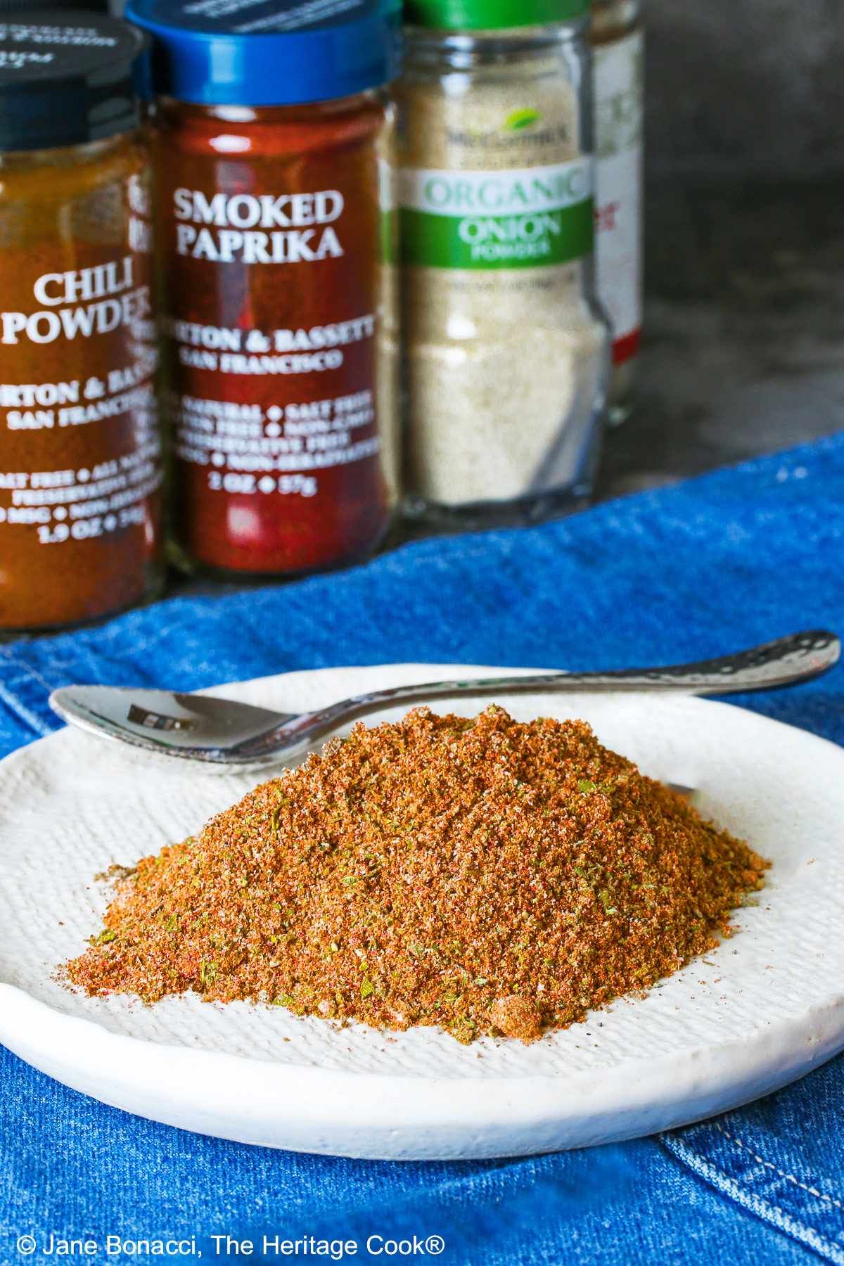 Homemade Taco Seasoning piled on a small white plate with bottles of the included seasonings behind, sitting on a blue cloth with a small spoon next to the taco seasoning © 2025 Jane Bonacci, The Heritage Cook.
