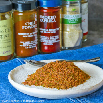 Homemade Taco Seasoning piled on a small white plate with bottles of the included seasonings behind, sitting on a blue cloth with a small spoon next to the taco seasoning © 2025 Jane Bonacci, The Heritage Cook.