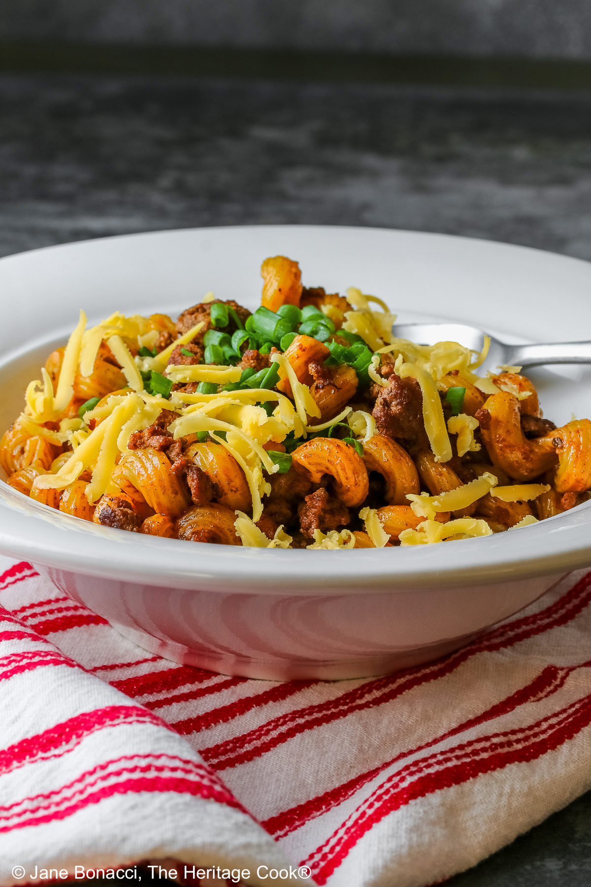 Tex-Mex Chili Mac made in the Instant Pot scooped into a white bowl with a wide rim, topped with chopped chives and extra shredded cheese, sitting on a red and white striped cloth © 2025 Jane Bonacci, The Heritage Cook. 