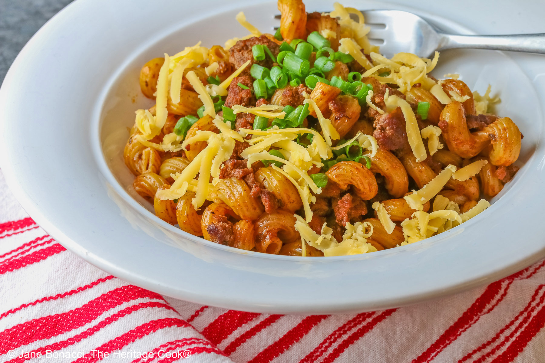 Tex-Mex Chili Mac made in the Instant Pot scooped into a white bowl with a wide rim, topped with chopped chives and extra shredded cheese, sitting on a red and white striped cloth © 2025 Jane Bonacci, The Heritage Cook. 