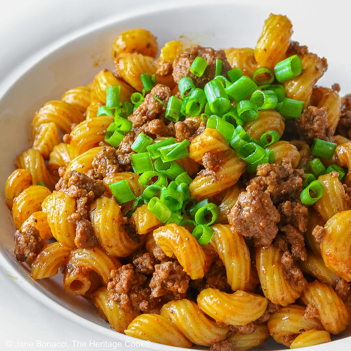 Ready for final sprinkling of shredded cheese; Tex-Mex Chili Mac made in the Instant Pot scooped into a white bowl with a wide rim, topped with chopped chives and extra shredded cheese, sitting on a red and white striped cloth © 2025 Jane Bonacci, The Heritage Cook. 