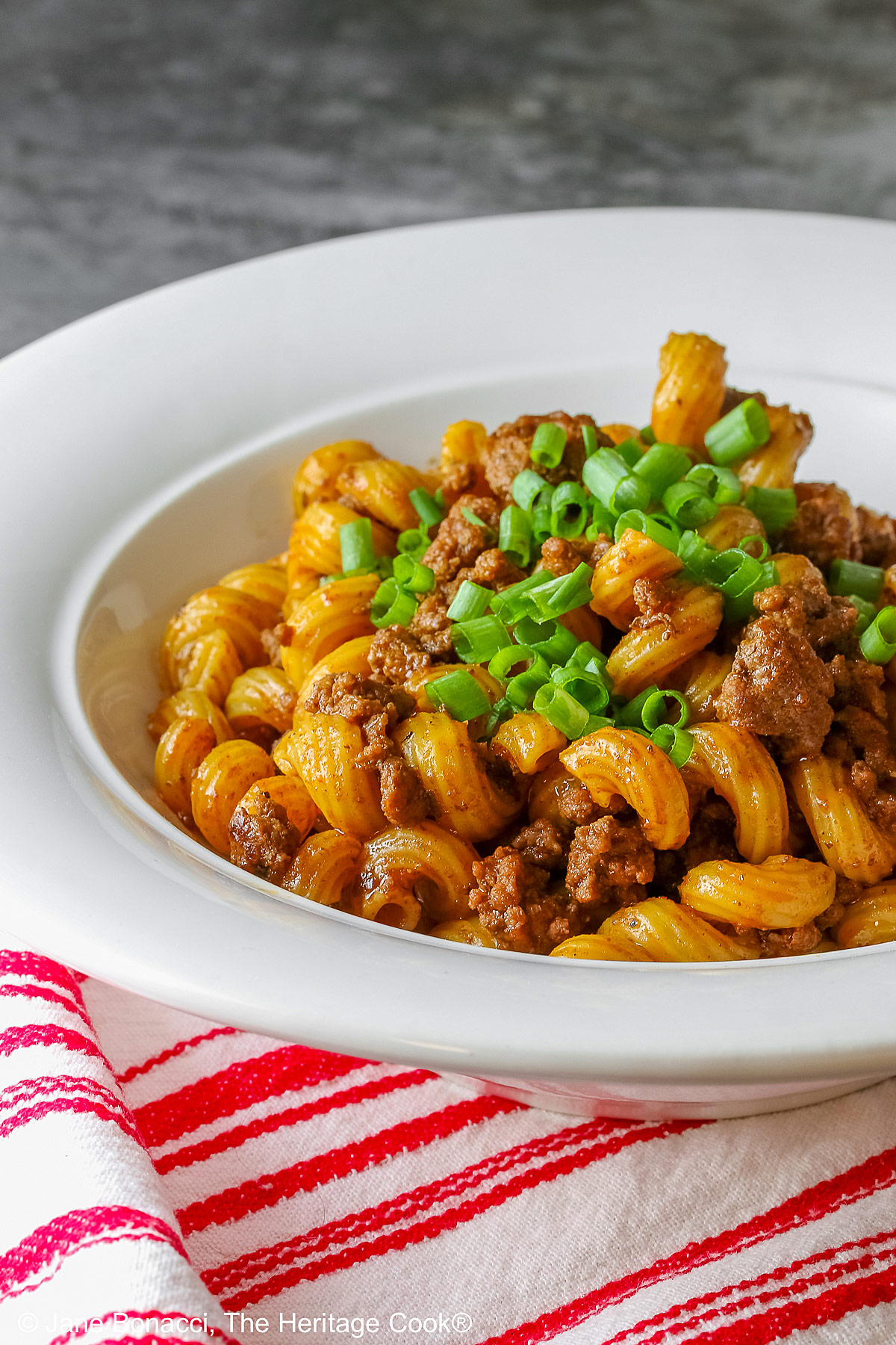 Tex-Mex Chili Mac made in the Instant Pot scooped into a white bowl with a wide rim, topped with chopped chives and extra shredded cheese, sitting on a red and white striped cloth © 2025 Jane Bonacci, The Heritage Cook. 