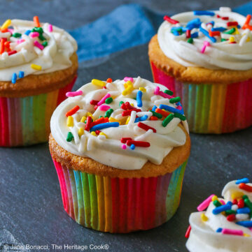 Five Vanilla Cupcakes with White Chocolate Frosting baked in vivid rainbow colored paper liners topped with creamy, fluffy white frosting and brightly colored festive sprinkles sitting on a black slate rectangle with a blue cloth behind © 2025 Jane Bonacci, The Heritage Cook.