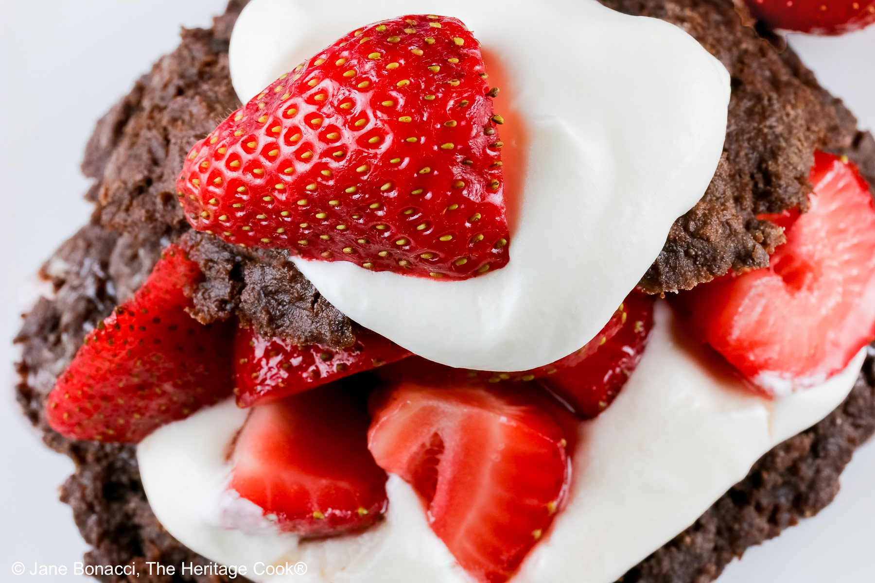 Chocolate shortcake, split and filled with whipped cream and sugared berries, covered with the chocolate top, a dollop of whipped cream, and more berries on a white plate with a blue background © 2025 Jane Bonacci, The Heritage Cook.