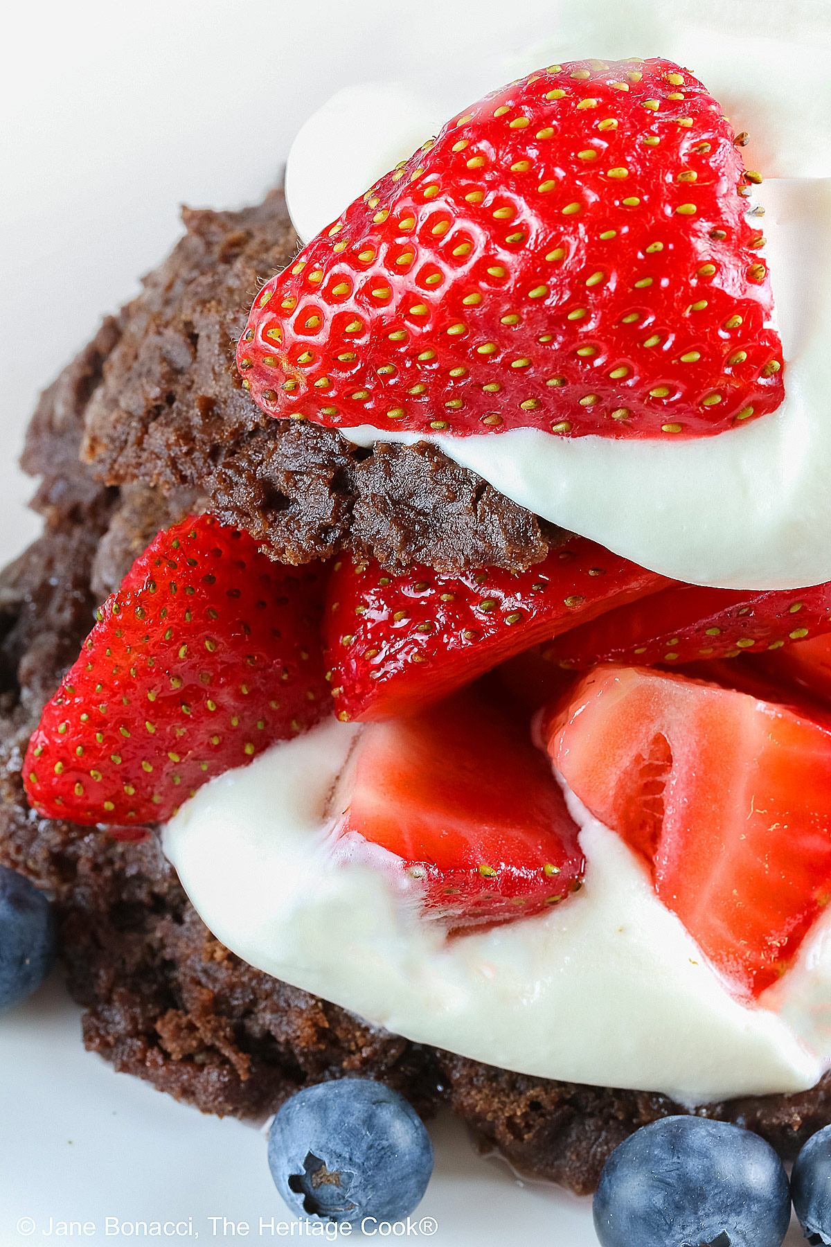 Chocolate shortcake, split and filled with whipped cream and sugared berries, covered with the chocolate top, a dollop of whipped cream, and more berries on a white plate with a blue background © 2025 Jane Bonacci, The Heritage Cook.