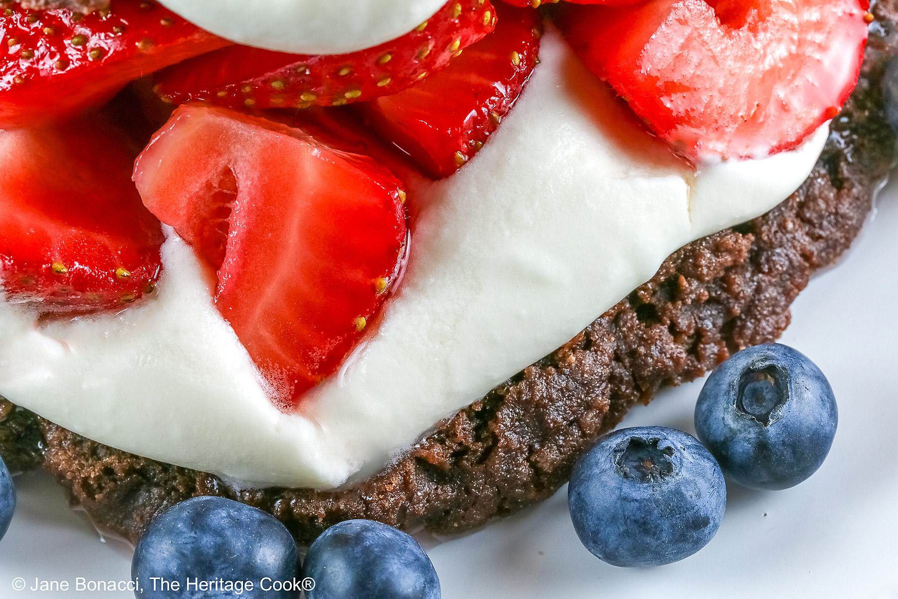 Chocolate shortcake, split and filled with whipped cream and sugared berries, covered with the chocolate top, a dollop of whipped cream, and more berries on a white plate with a blue background © 2025 Jane Bonacci, The Heritage Cook.