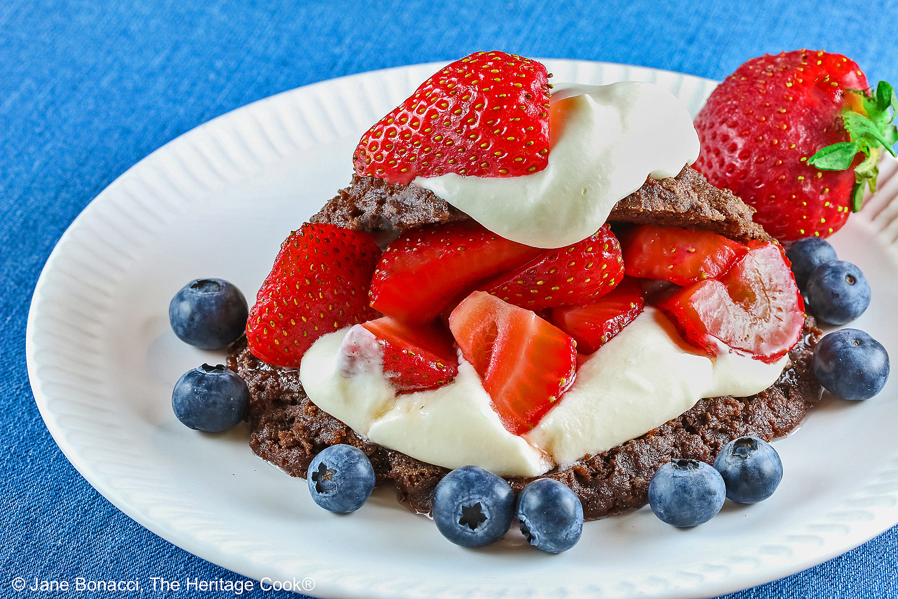 Chocolate shortcake, split and filled with whipped cream and sugared berries, covered with the chocolate top, a dollop of whipped cream, and more berries on a white plate with a blue background © 2025 Jane Bonacci, The Heritage Cook.