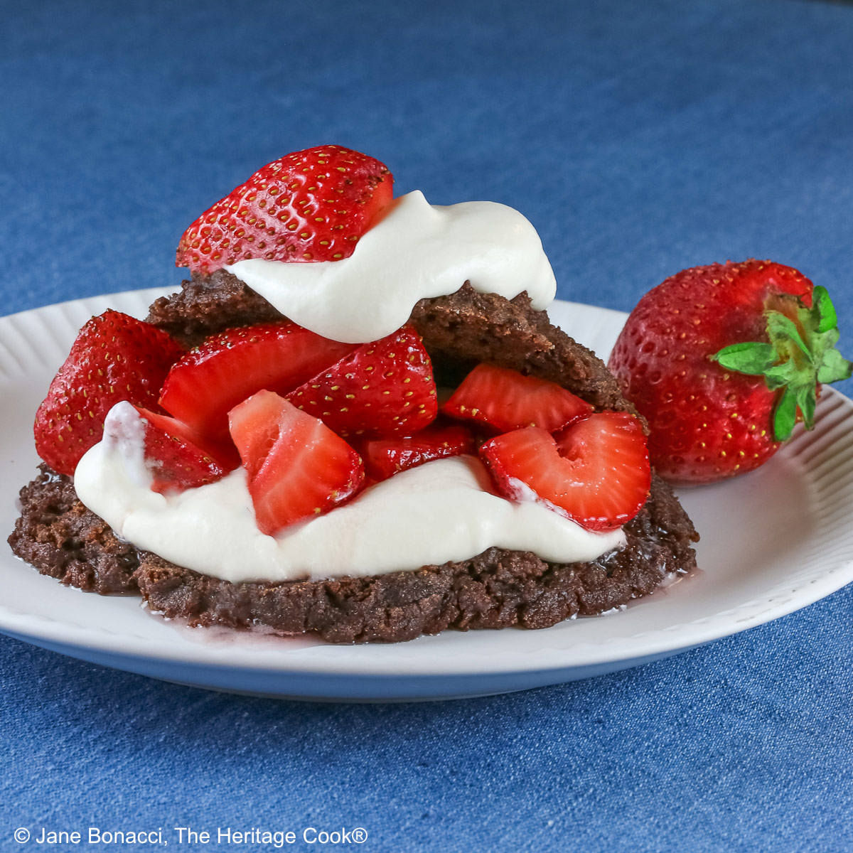Chocolate shortcake, split and filled with whipped cream and sugared berries, covered with the chocolate top, a dollop of whipped cream, and more berries on a white plate with a blue background © 2025 Jane Bonacci, The Heritage Cook.