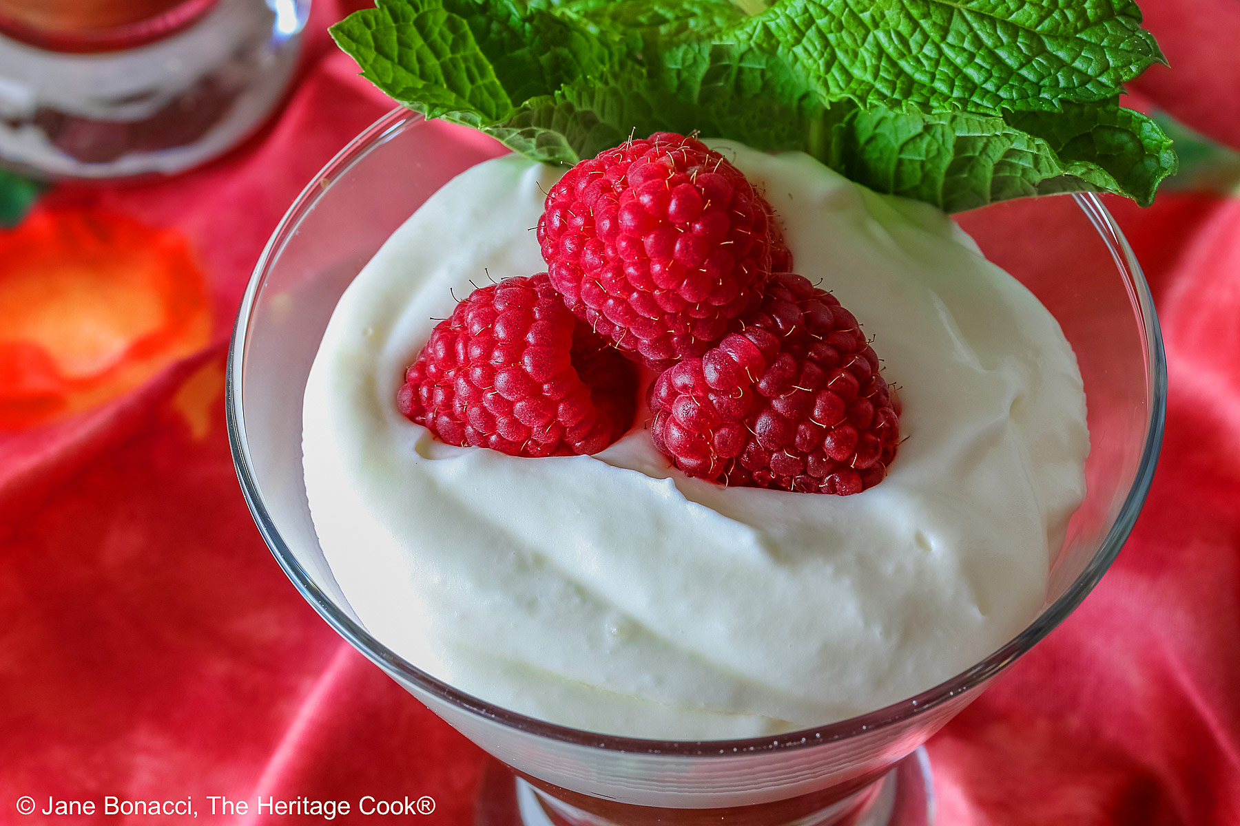 Two glass dishes with Egg-Free Bourbon Chocolate Mousse topped with freshly whipped cream and garnished with fresh raspberries and a sprig of mint, sitting on a deep pink/red cloth © 2025 Jane Bonacci, The Heritage Cook. 
