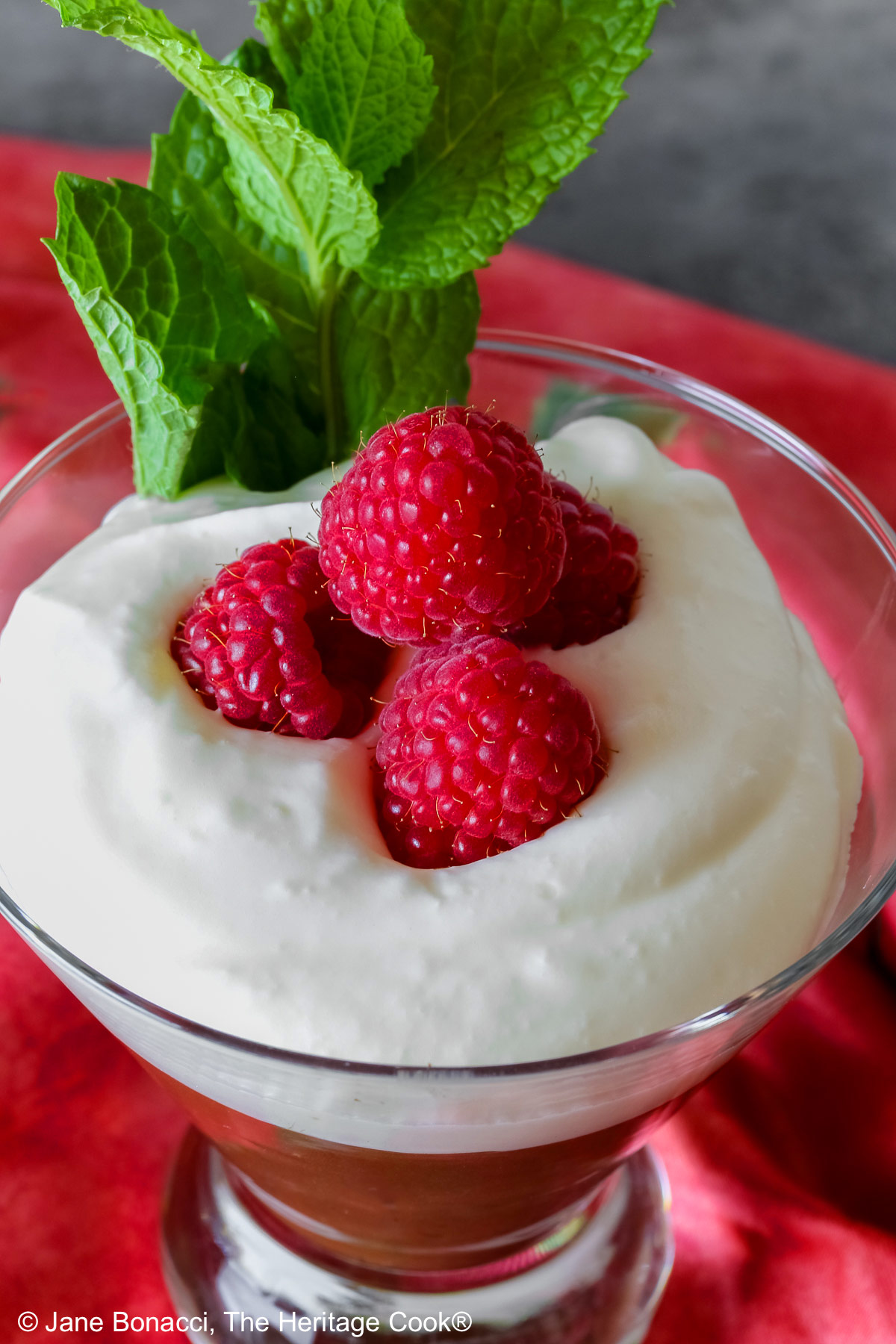 Two glass dishes with Egg-Free Bourbon Chocolate Mousse topped with freshly whipped cream and garnished with fresh raspberries and a sprig of mint, sitting on a deep pink/red cloth © 2025 Jane Bonacci, The Heritage Cook. 