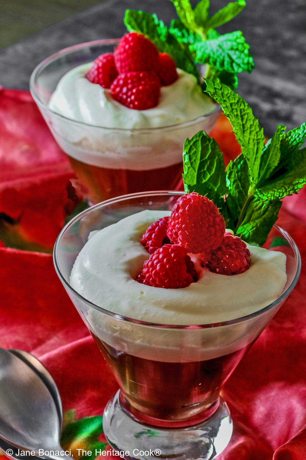 Two glass dishes with Egg-Free Bourbon Chocolate Mousse topped with freshly whipped cream and garnished with fresh raspberries and a sprig of mint, sitting on a deep pink/red cloth © 2025 Jane Bonacci, The Heritage Cook. 