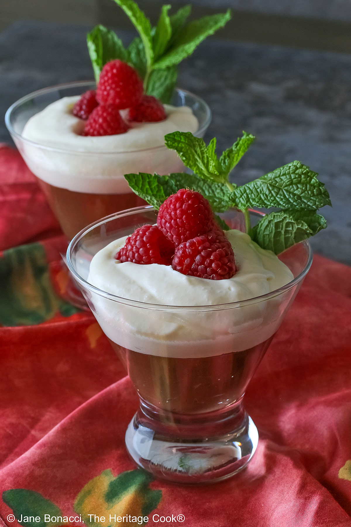 Two glass dishes with Egg-Free Bourbon Chocolate Mousse topped with freshly whipped cream and garnished with fresh raspberries and a sprig of mint, sitting on a deep pink/red cloth © 2025 Jane Bonacci, The Heritage Cook. 