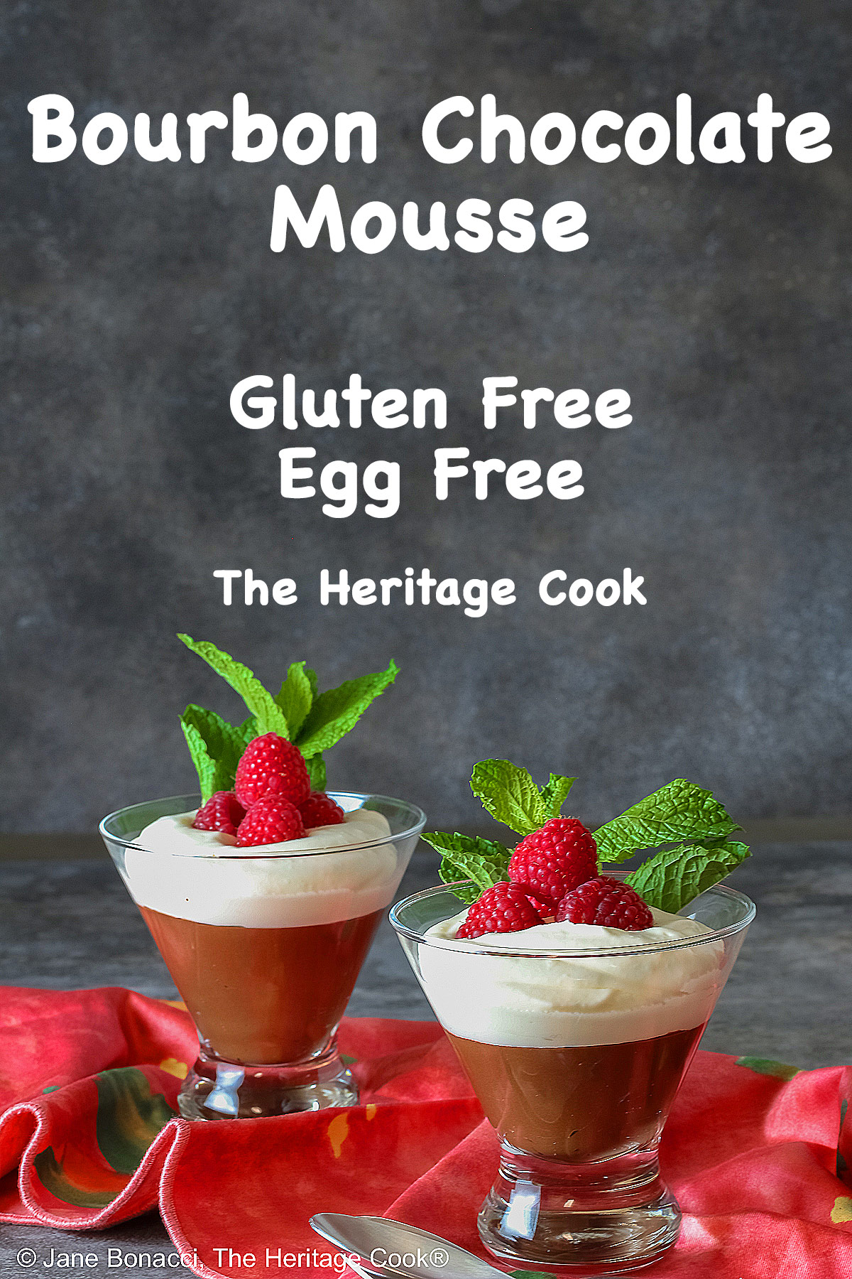 Two glass dishes with Egg-Free Bourbon Chocolate Mousse topped with freshly whipped cream and garnished with fresh raspberries and a sprig of mint, sitting on a deep pink/red cloth © 2025 Jane Bonacci, The Heritage Cook. 