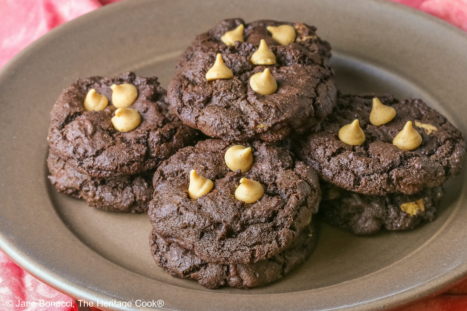 Four stacks of black Double Chocolate Peanut Butter Cookies with light peanut butter chips on top on a deep brown ceramic plate sitting on a bright pink cloth © 2025 Jane Bonacci, The Heritage Cook.