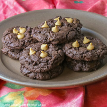 Four stacks of black Double Chocolate Peanut Butter Cookies with light peanut butter chips on top on a deep brown ceramic plate sitting on a bright pink cloth © 2025 Jane Bonacci, The Heritage Cook.
