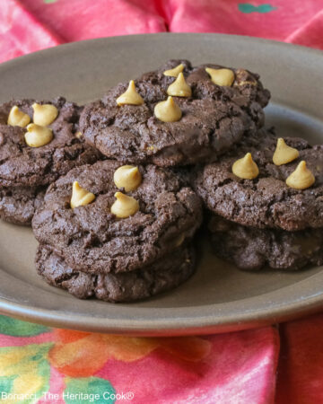 Four stacks of black Double Chocolate Peanut Butter Cookies with light peanut butter chips on top on a deep brown ceramic plate sitting on a bright pink cloth © 2025 Jane Bonacci, The Heritage Cook.