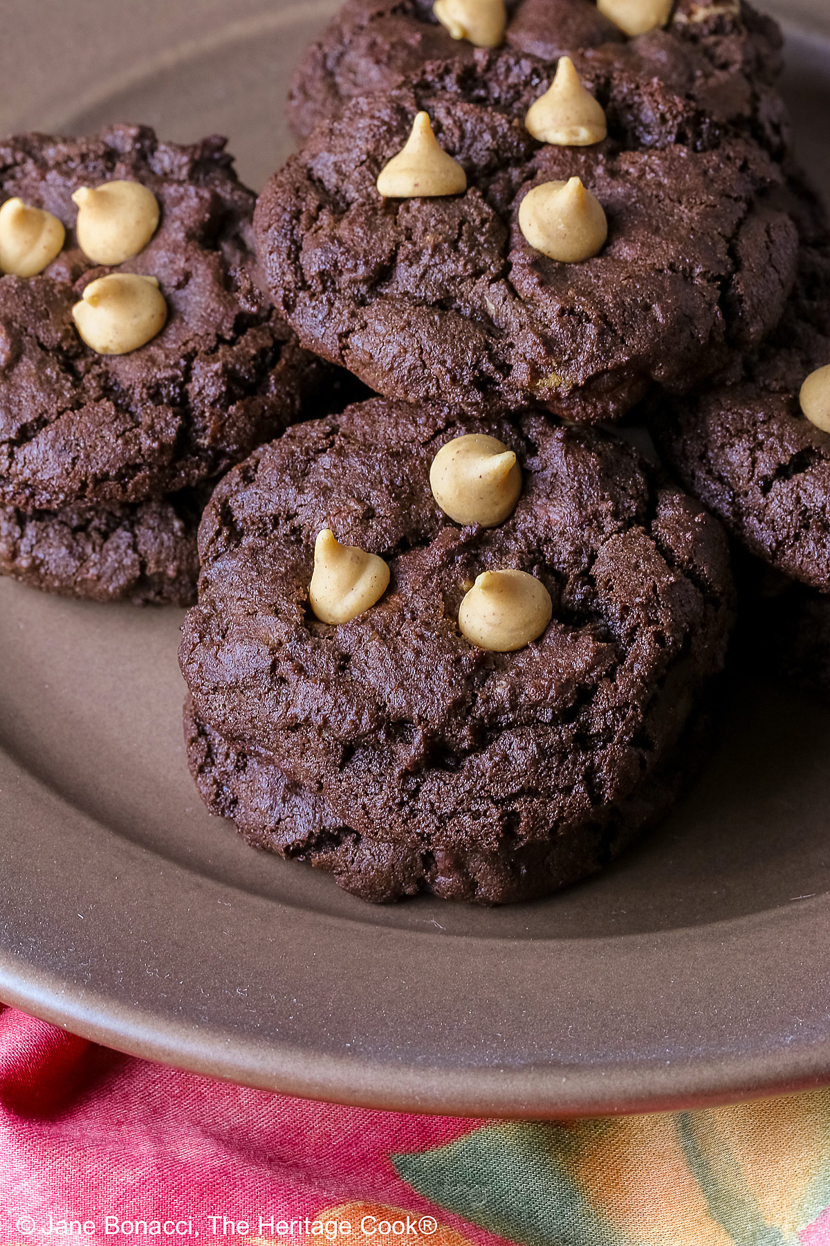 Four stacks of black Double Chocolate Peanut Butter Cookies with light peanut butter chips on top on a deep brown ceramic plate sitting on a bright pink cloth © 2025 Jane Bonacci, The Heritage Cook.