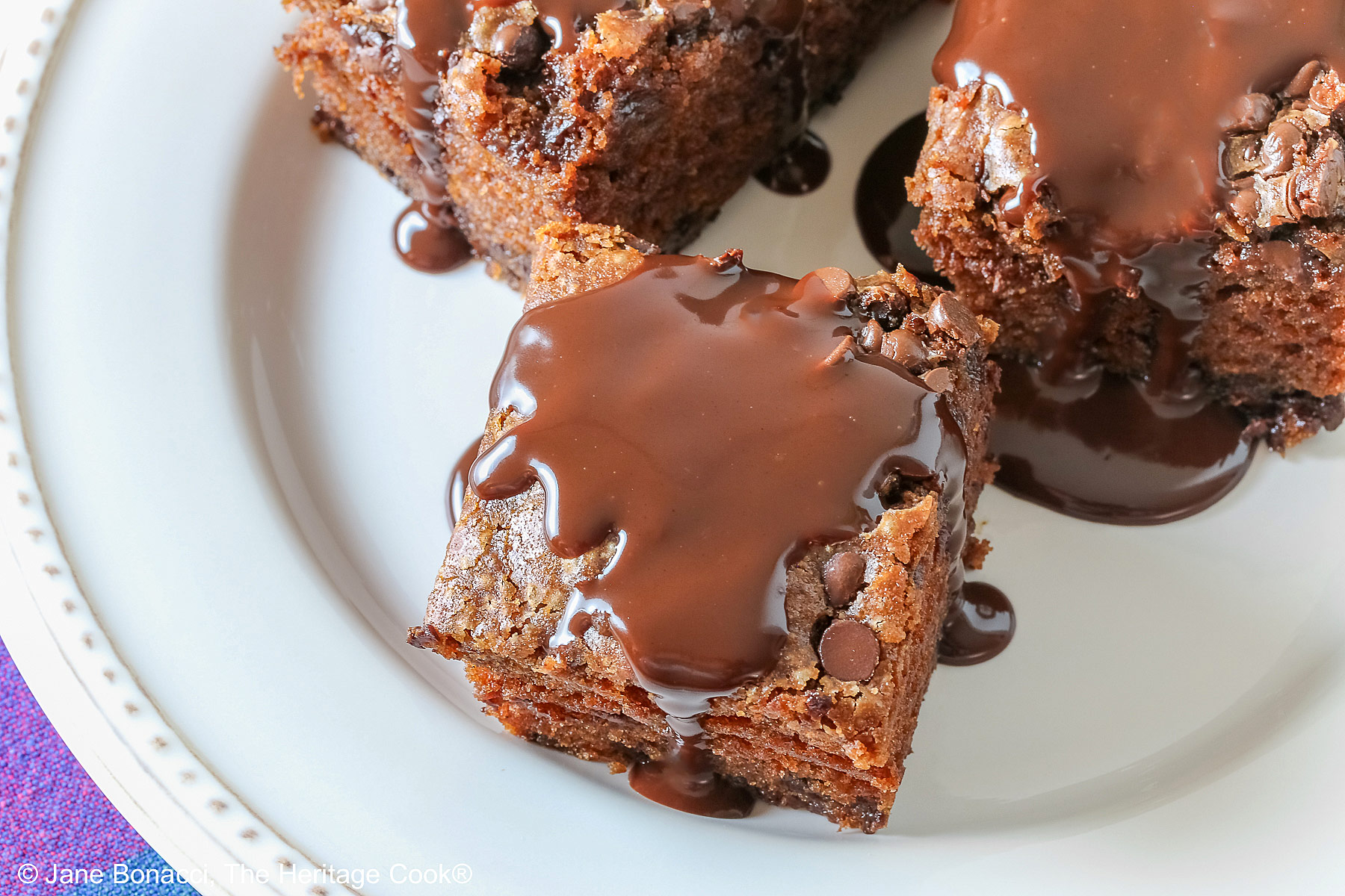 3 squares of Egg-Free Chocolate, Chocolate Chip Cake on a white plate with a couple of sprigs of mint, sitting on a brightly colored plaid cloth © 2025 Jane Bonacci, The Heritage Cook. 