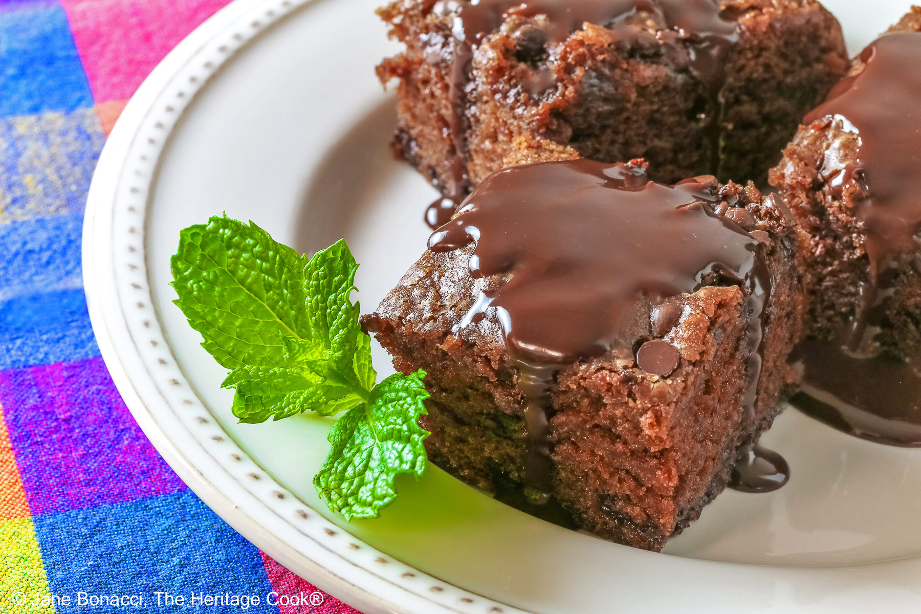 3 squares of Egg-Free Chocolate, Chocolate Chip Cake on a white plate with a couple of sprigs of mint, sitting on a brightly colored plaid cloth © 2025 Jane Bonacci, The Heritage Cook. 