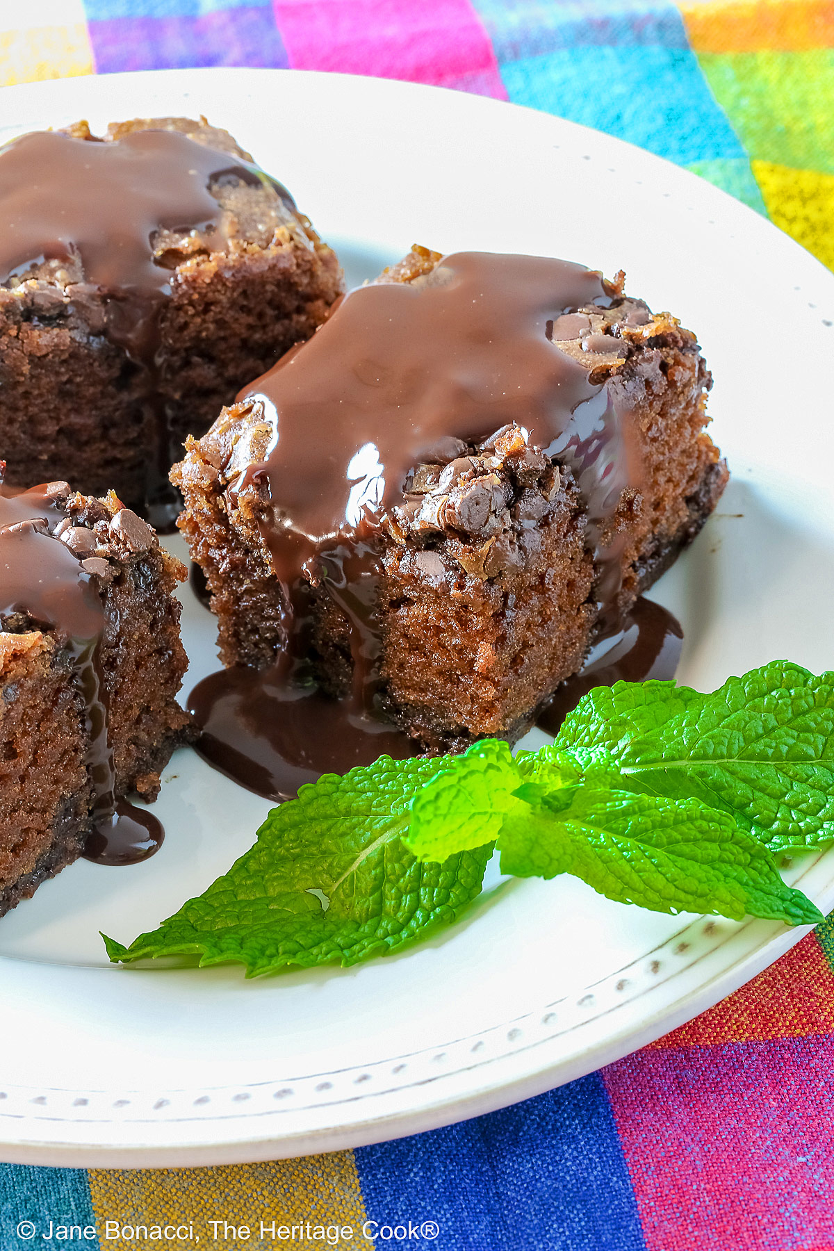 3 squares of Egg-Free Chocolate, Chocolate Chip Cake on a white plate with a couple of sprigs of mint, sitting on a brightly colored plaid cloth © 2025 Jane Bonacci, The Heritage Cook. 