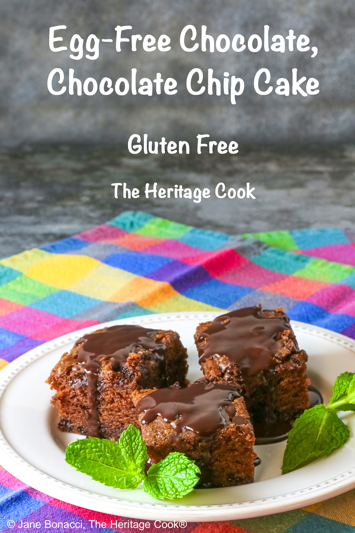 3 squares of Egg-Free Chocolate, Chocolate Chip Cake on a white plate with a couple of sprigs of mint, sitting on a brightly colored plaid cloth © 2025 Jane Bonacci, The Heritage Cook.