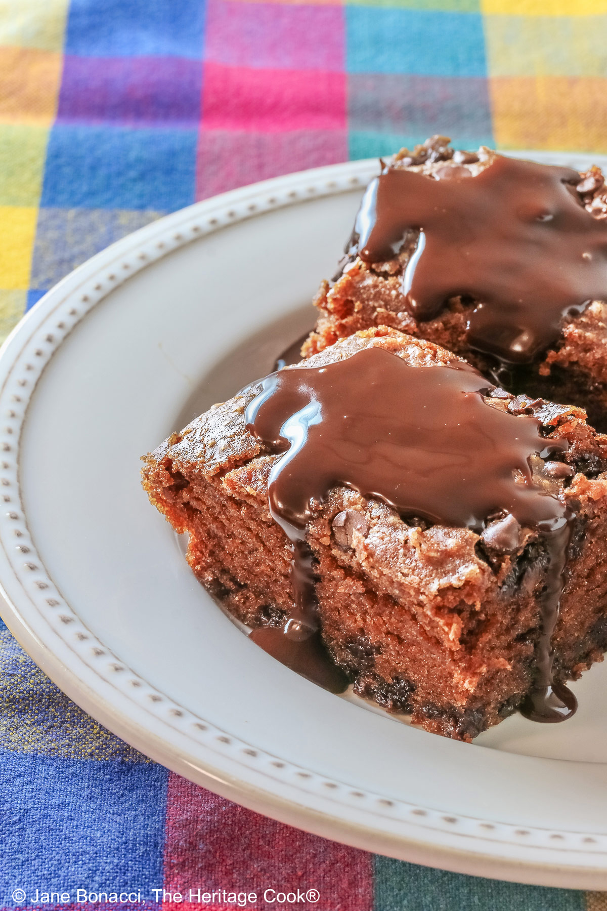3 squares of Egg-Free Chocolate, Chocolate Chip Cake on a white plate with a couple of sprigs of mint, sitting on a brightly colored plaid cloth © 2025 Jane Bonacci, The Heritage Cook. 