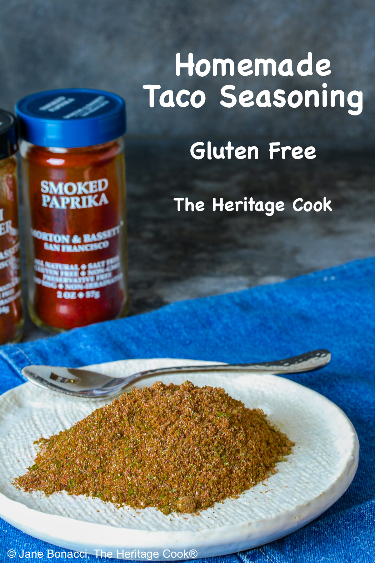 Homemade Taco Seasoning piled on a small white plate with bottles of the included seasonings behind, sitting on a blue cloth with a small spoon next to the taco seasoning © 2025 Jane Bonacci, The Heritage Cook. 