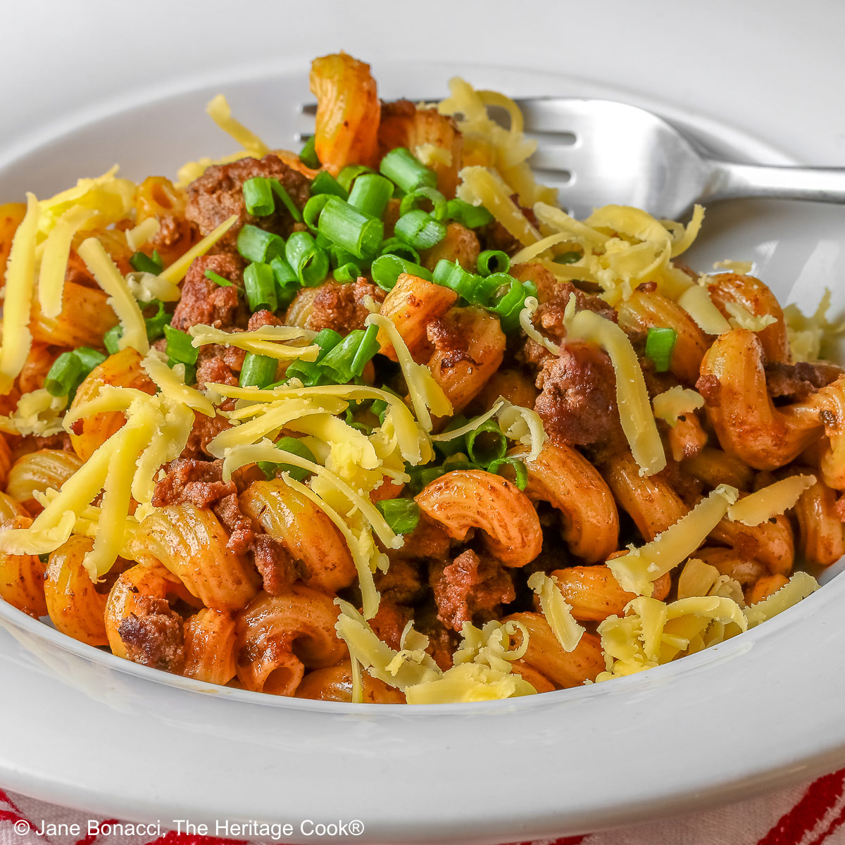 Tex-Mex Chili Mac made in the Instant Pot scooped into a white bowl with a wide rim, topped with chopped chives and extra shredded cheese, sitting on a red and white striped cloth © 2025 Jane Bonacci, The Heritage Cook.