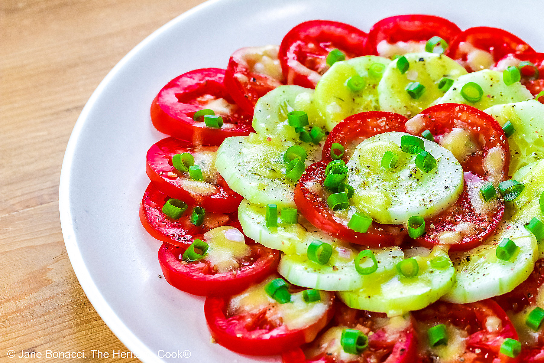 Concentric circles of tomatoes and cucumbers, each ring alternating between the two vegetables, drizzled with homemade vinaigrette and sprinkled with chopped green onions on a white plate © 2025 Jane Bonacci, The Heritage Cook. 