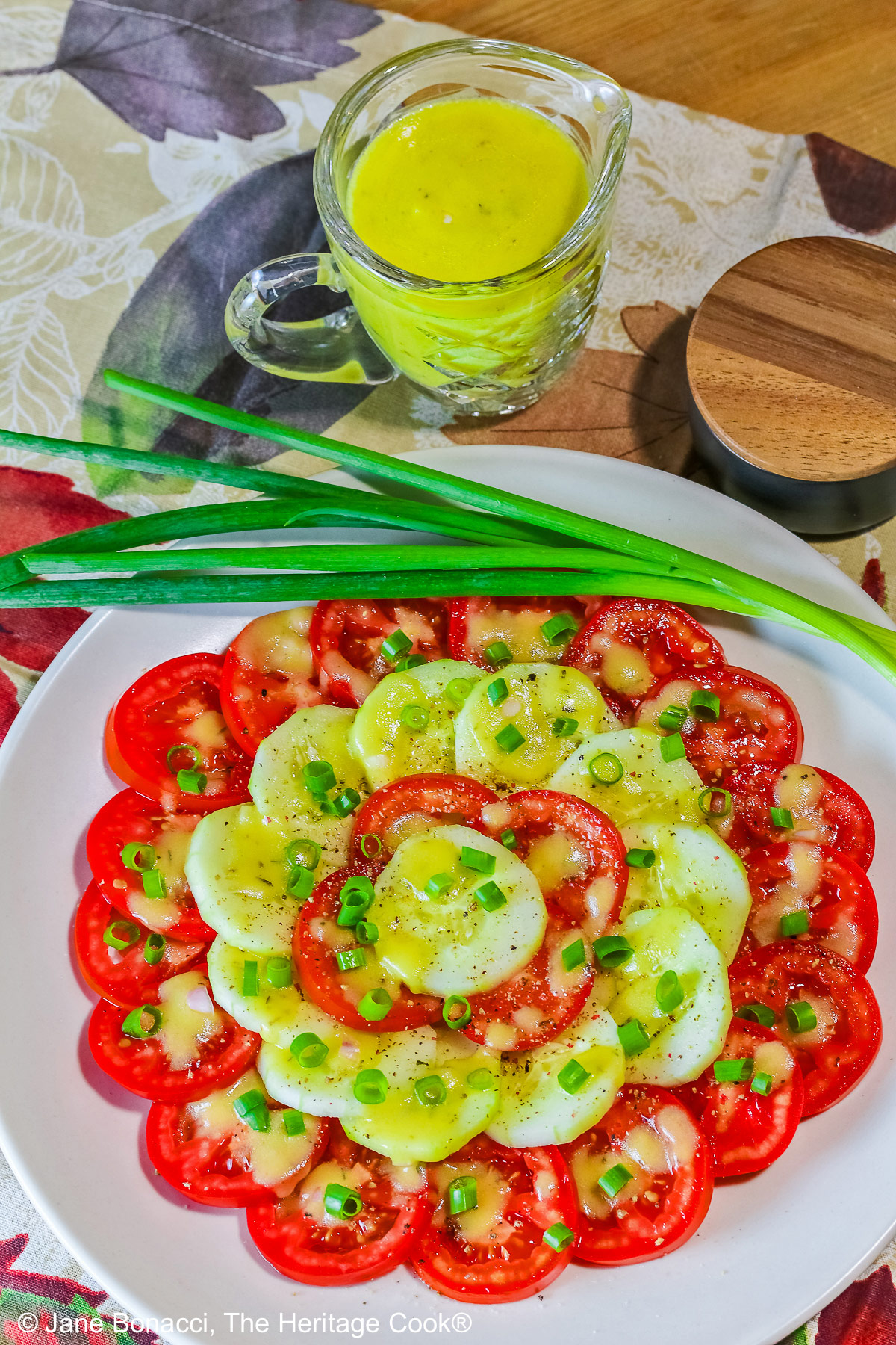 Concentric circles of tomatoes and cucumbers, each ring alternating between the two vegetables, drizzled with homemade vinaigrette and sprinkled with chopped green onions on a white plate © 2025 Jane Bonacci, The Heritage Cook. 