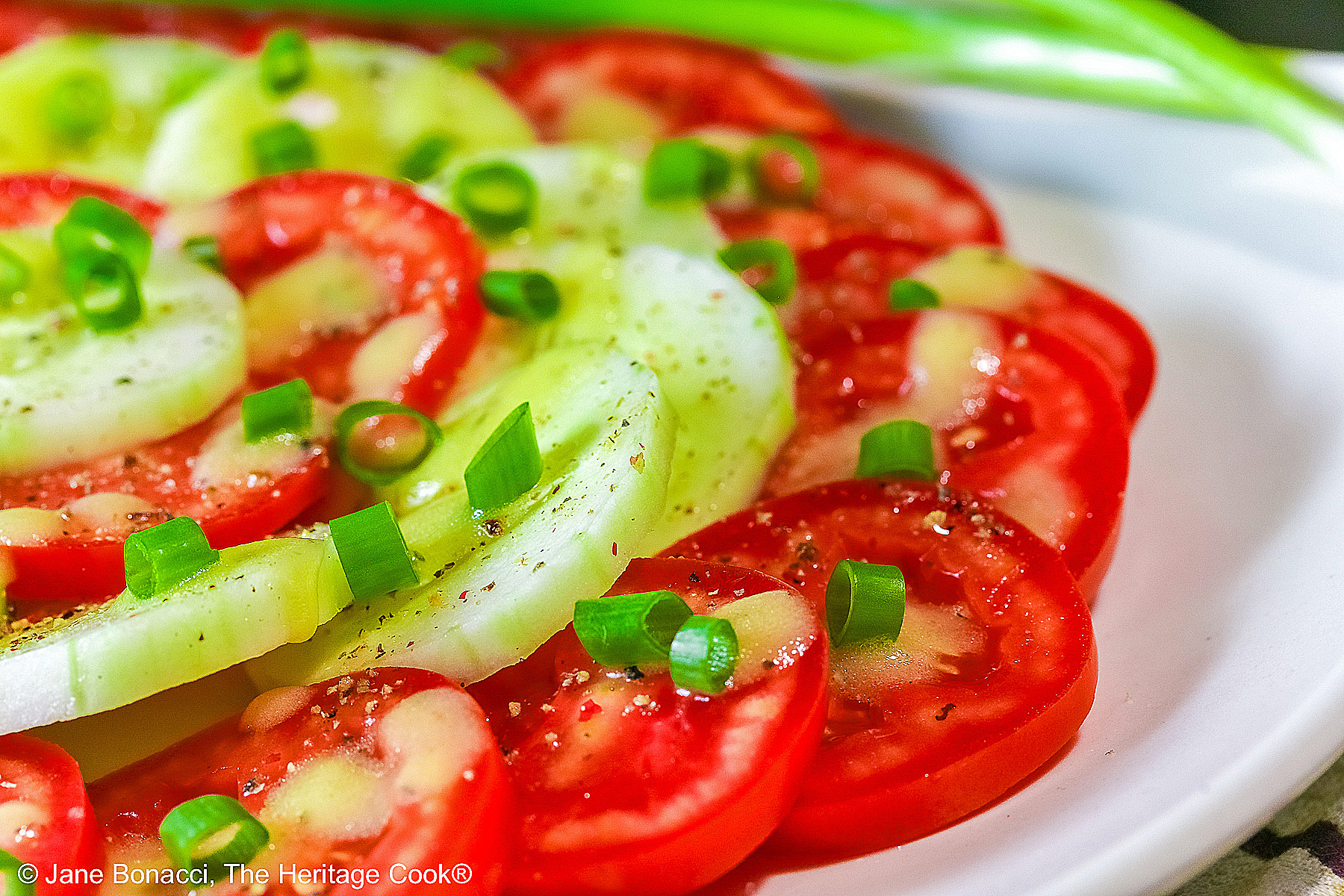 Concentric circles of tomatoes and cucumbers, each ring alternating between the two vegetables, drizzled with homemade vinaigrette and sprinkled with chopped green onions on a white plate © 2025 Jane Bonacci, The Heritage Cook. 