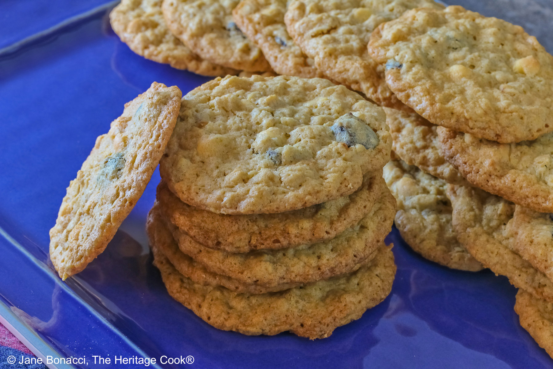 Elongated pile of White Chocolate Oatmeal Cherry cookies in the back with a stack of cookies in front and a single cookie propped against the stack on a blue rectangular platter with a multi-color cloth in front © 2025 Jane Bonacci, The Heritage Cook.