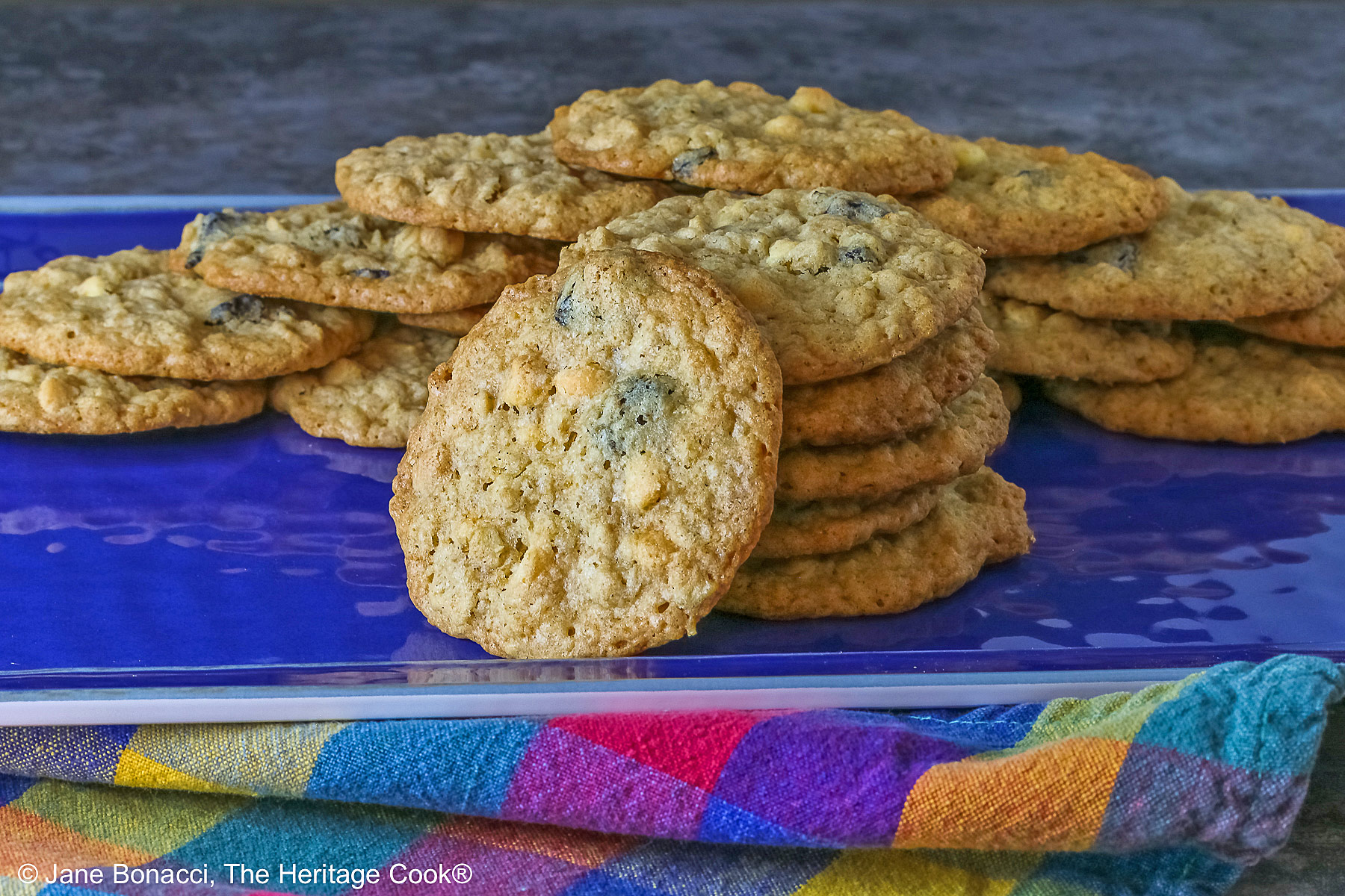 Elongated pile of White Chocolate Oatmeal Cherry cookies in the back with a stack of cookies in front and a single cookie propped against the stack on a blue rectangular platter with a multi-color cloth in front © 2025 Jane Bonacci, The Heritage Cook.