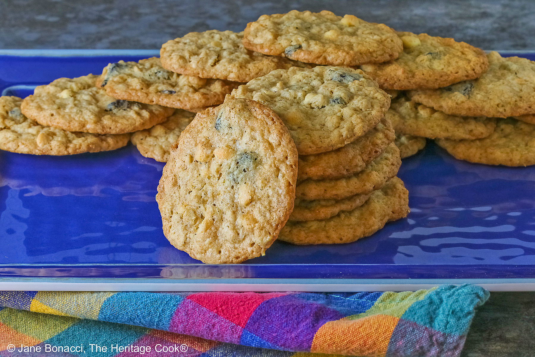 Elongated pile of White Chocolate Oatmeal Cherry cookies in the back with a stack of cookies in front and a single cookie propped against the stack on a blue rectangular platter with a multi-color cloth in front © 2025 Jane Bonacci, The Heritage Cook.