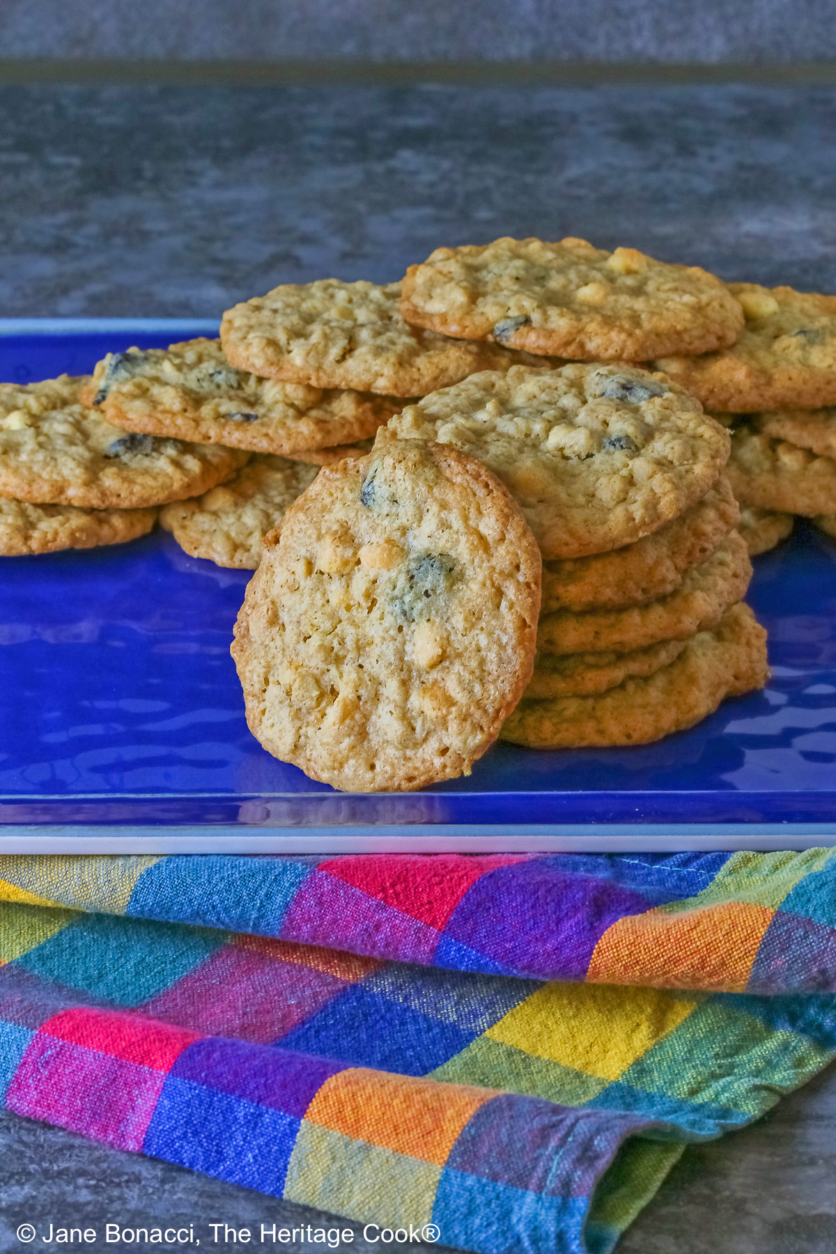 Elongated pile of White Chocolate Oatmeal Cherry cookies in the back with a stack of cookies in front and a single cookie propped against the stack on a blue rectangular platter with a multi-color cloth in front © 2025 Jane Bonacci, The Heritage Cook.