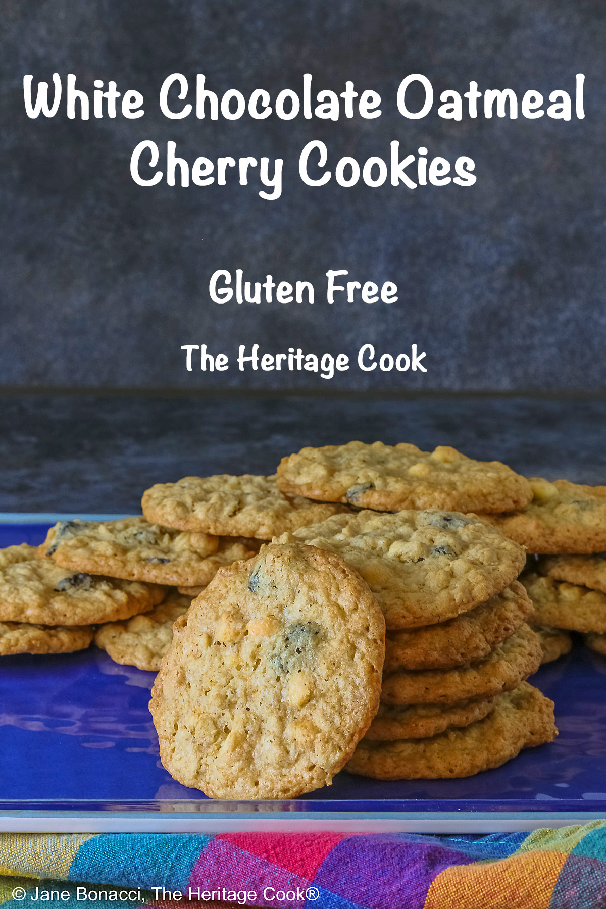 Elongated pile of White Chocolate Oatmeal Cherry cookies in the back with a stack of cookies in front and a single cookie propped against the stack on a blue rectangular platter with a multi-color cloth in front © 2025 Jane Bonacci, The Heritage Cook.