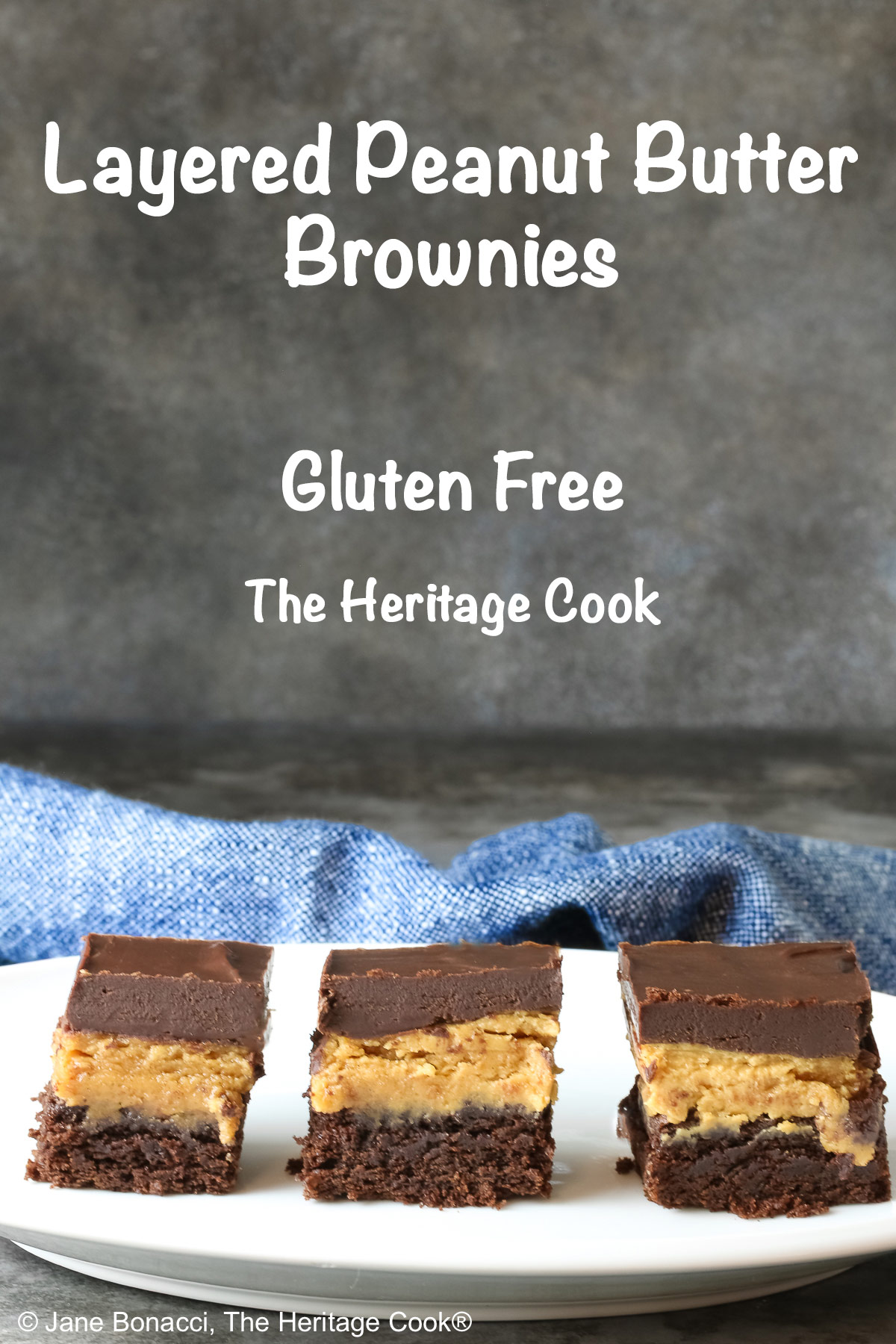 3 Layered Peanut Butter Brownies on an oval white platter with a blue cloth behind and a gray mottled background; brownie on the bottom, peanut butter layer in the middle and finished with a thick ganache chocolate layer on top ©2025 Jane Bonacci, The Heritage Cook. 
