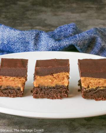 3 Layered Peanut Butter Brownies on an oval white platter with a blue cloth behind and a gray mottled background; brownie on the bottom, peanut butter layer in the middle and finished with a thick ganache chocolate layer on top ©2025 Jane Bonacci, The Heritage Cook.