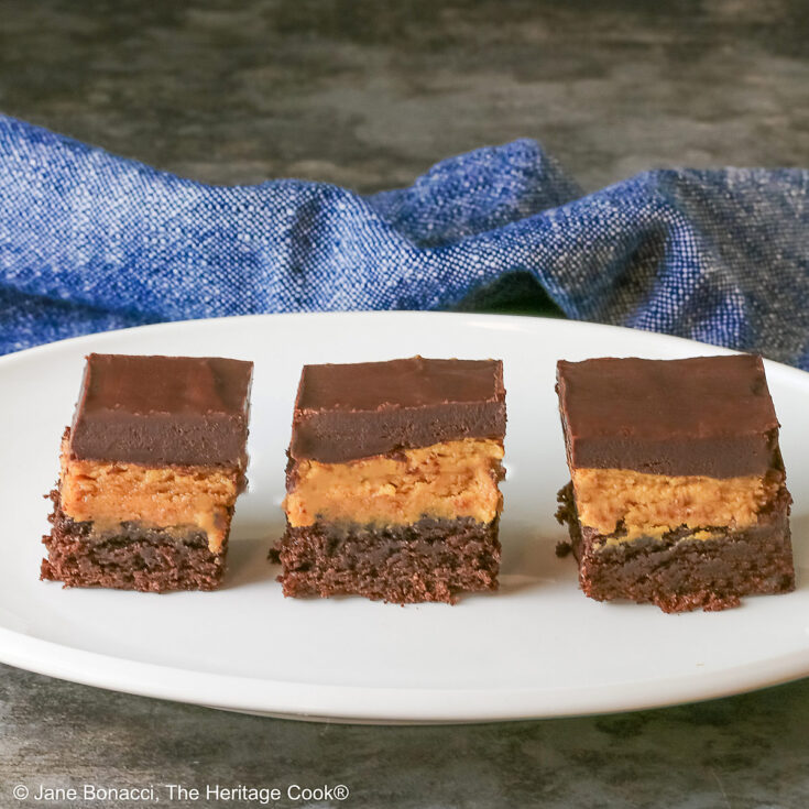 3 Layered Peanut Butter Brownies on an oval white platter with a blue cloth behind and a gray mottled background; brownie on the bottom, peanut butter layer in the middle and finished with a thick ganache chocolate layer on top ©2025 Jane Bonacci, The Heritage Cook.
