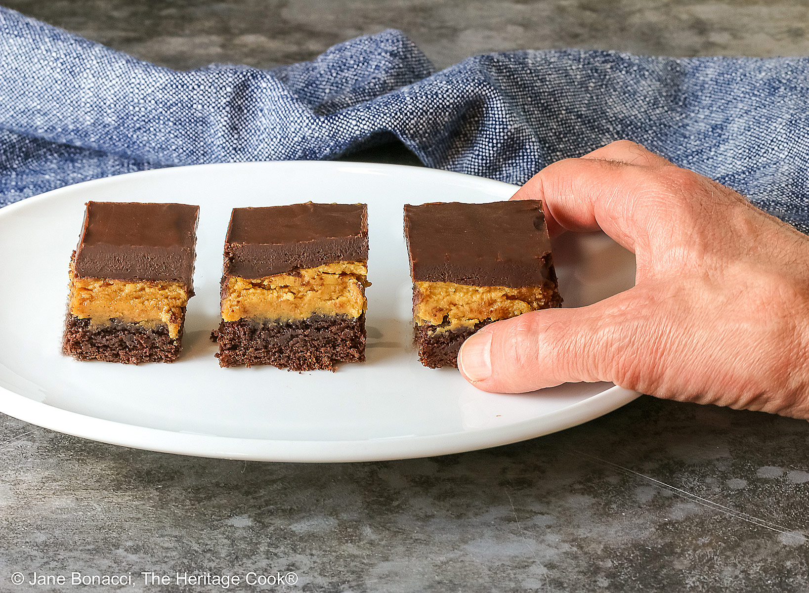 Hand reaching to grab a brownie; 3 Layered Peanut Butter Brownies on an oval white platter with a blue cloth behind and a gray mottled background; brownie on the bottom, peanut butter layer in the middle and finished with a thick ganache chocolate layer on top ©2025 Jane Bonacci, The Heritage Cook. 